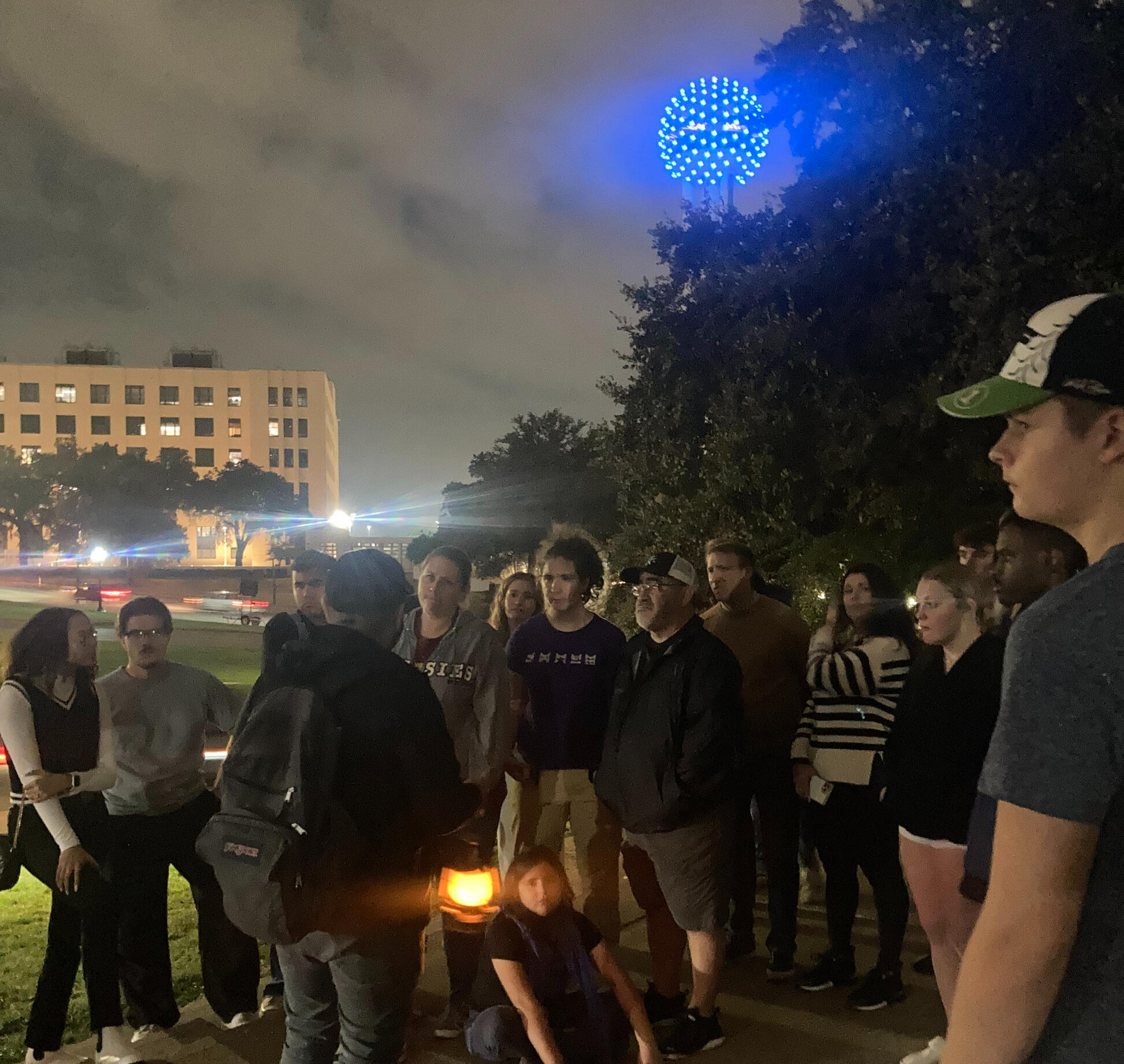 A photo from one of our true crime walking tours showing reunion tower lit in blue neon A photo from one of our true crime walking tours showing reunion tower at dusk lit in blue neon a crowd of people gather to listen to our guide tell his story